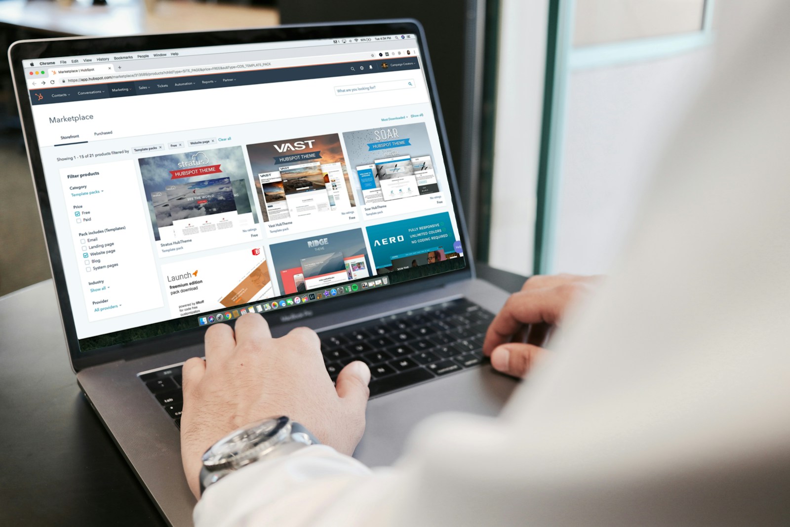 A person is using a laptop to browse an online marketplace featuring various digital products or themes. The screen displays a selection of software or website themes with descriptions and prices. The person seems to be in a well-lit room, sitting at a table.